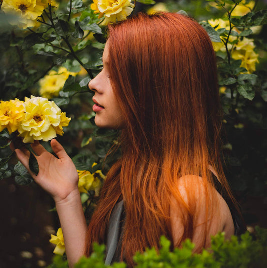 Woman with long, healthy auburn hair standing in a garden with yellow flowers, symbolizing natural shine and hair vitality.