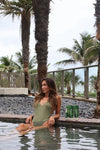 FUYA Haircare lifestyle – woman in green swimsuit sitting by the pool with FUYA Repair & Shine products and palm trees in the background